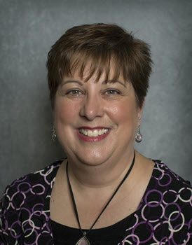 A photo of a woman with short brown hair wearing a patterned black blouse in front of a gray background.