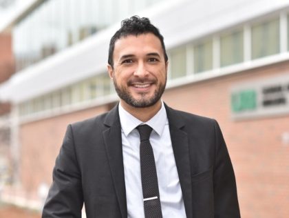 Photo of a smiling man with dark hair and a beard, wearing a suit.