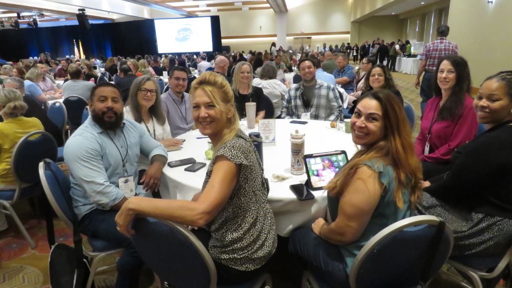 Large Conference room with round tables at Albuquerque conference, zoomed on one table of smiling participants.