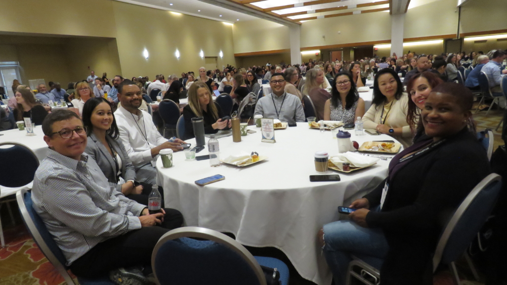 Large Conference room with round tables at Albuquerque conference, zoomed on one table of smiling participants.