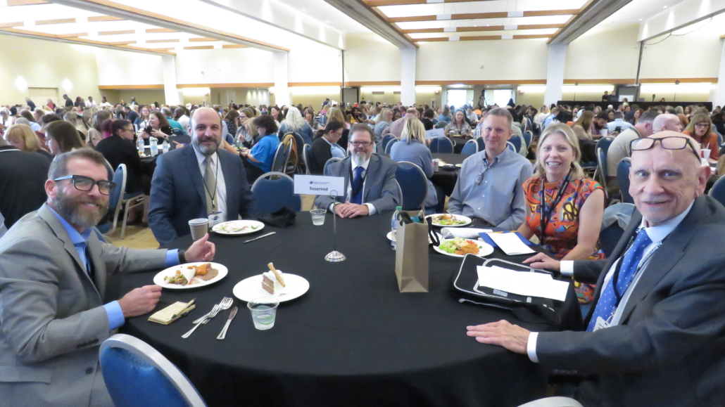 Large Conference room with round tables at Albuquerque conference, zoomed on one table of smiling participants.