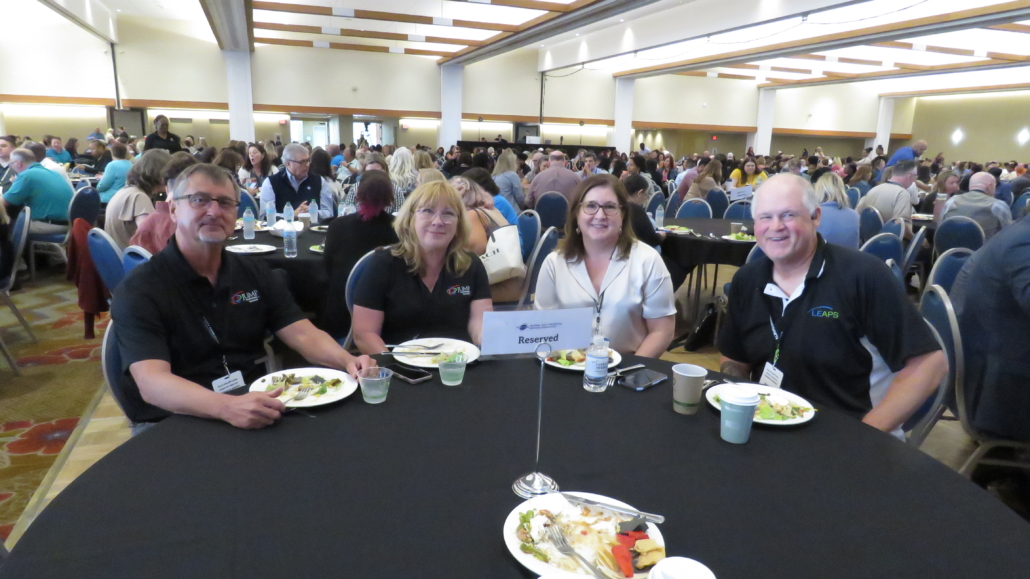 Large Conference room with round tables at the Annual Conference 2024, zoomed on a group at one table smiling.