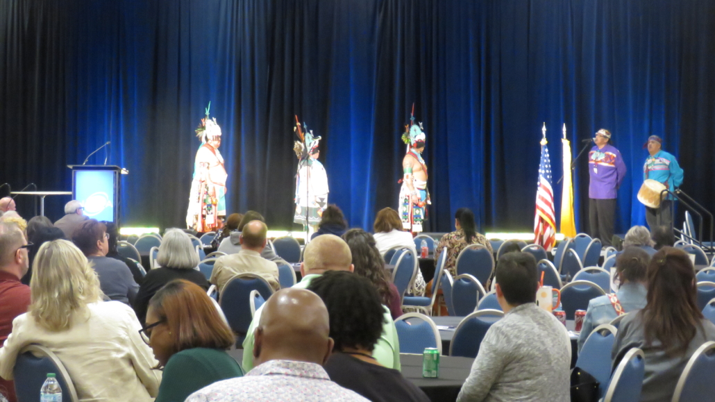 Five presenters on stage, one with a drum, and an audience watching.